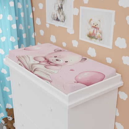 The baby changing pad cover featuring an adorable pink bear, with a soft pink background adorned with balloons, gift boxes, and heart motifs, placed in a room.