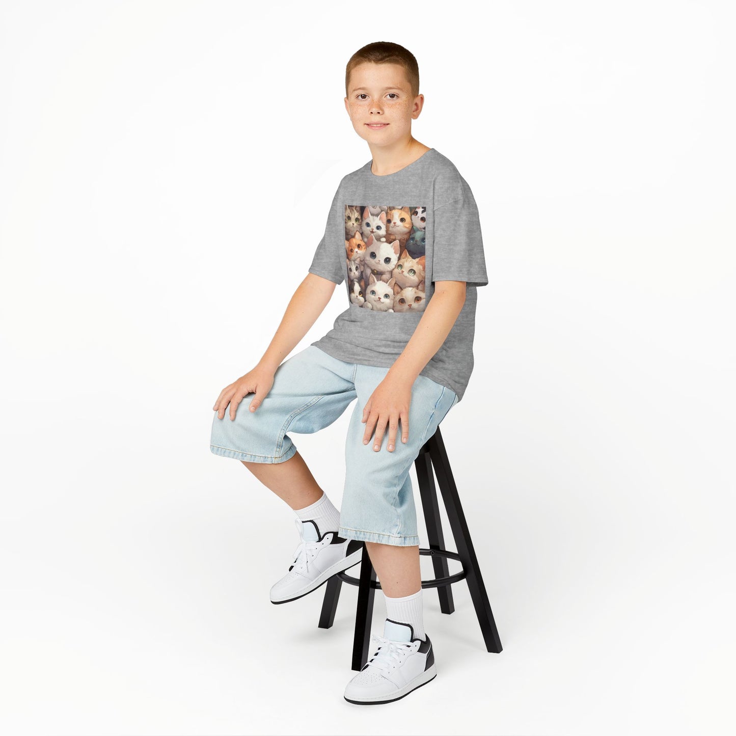 A young boy sitting on a chair, wearing a Sport Grey T-shirt featuring a charming design with an abundance of adorable cats.
