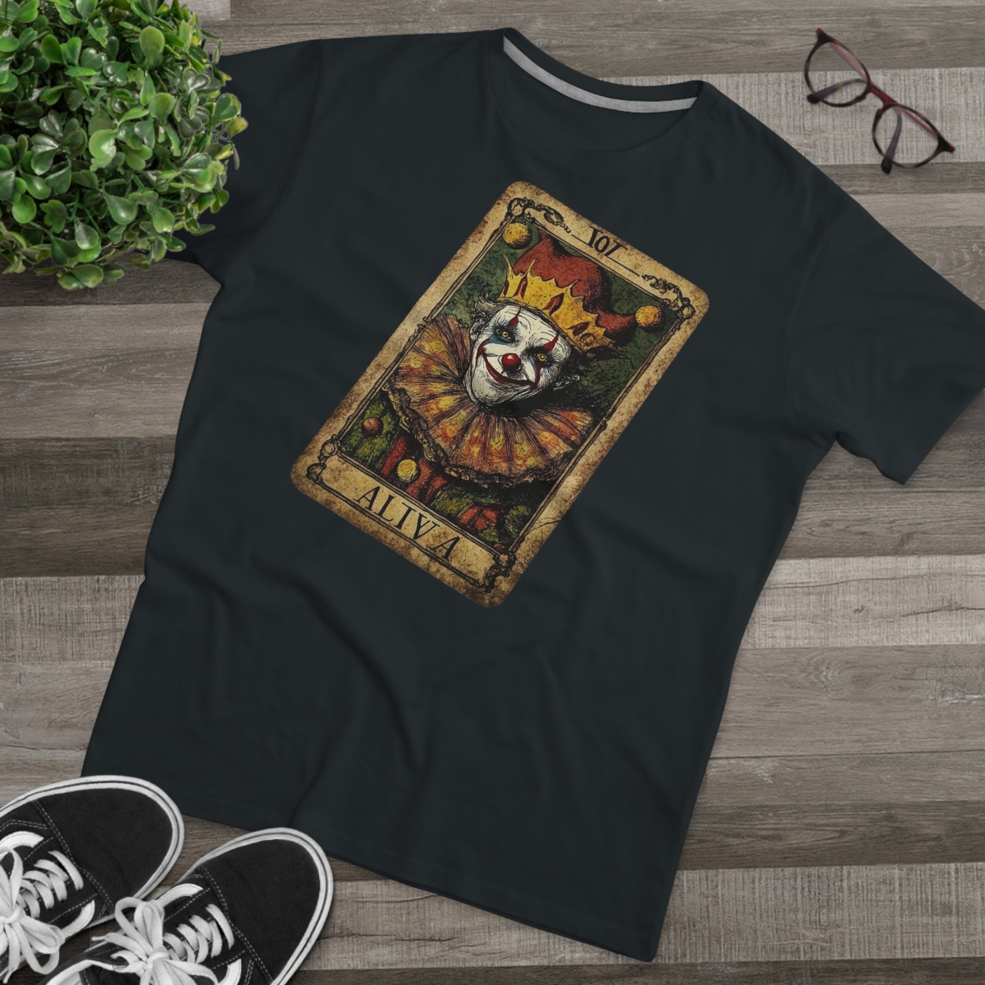 A Dark Black T-shirt with a Tarot card design featuring a clown against a dark grunge-style background, laid on the floor with glasses and shoes.