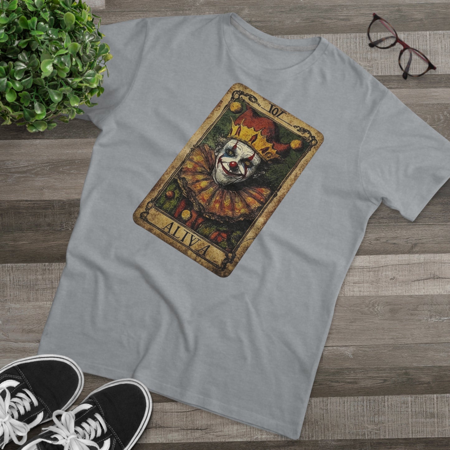 A Light Oxford T-shirt with a Tarot card design featuring a clown against a dark grunge-style background, laid on the floor with glasses and shoes.