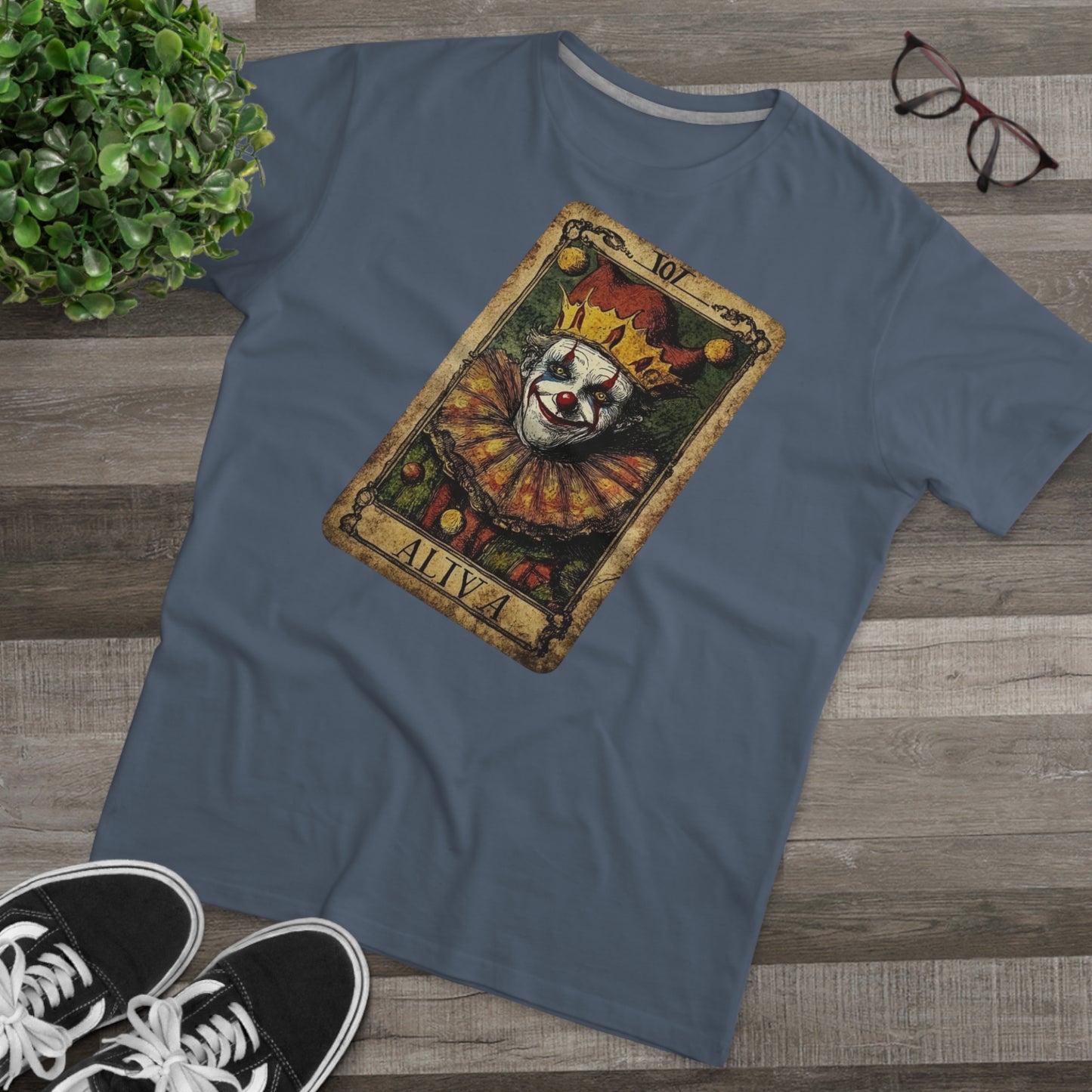 A Navy T-shirt with a Tarot card design featuring a clown against a dark grunge-style background, laid on the floor with glasses and shoes.