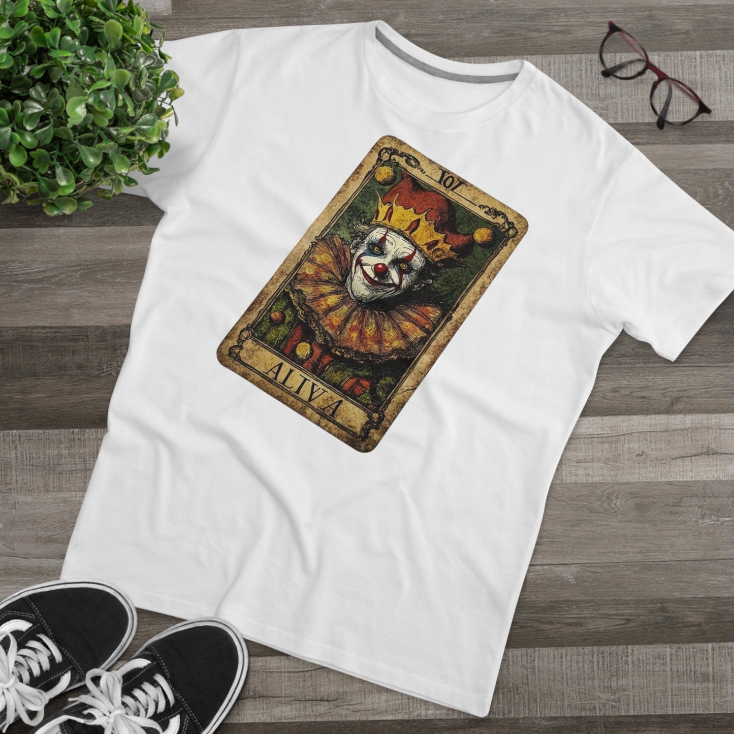 A White T-shirt with a Tarot card design featuring a clown against a dark grunge-style background, laid on the floor with glasses and shoes.