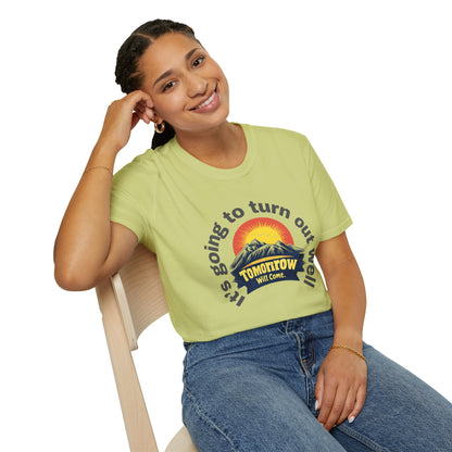 A woman sitting on a chair while wearing a pistachio-colored T-shirt designed with an uplifting message and a warm sunrise graphic.